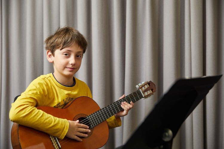A boy with brown hair and wearing a long sleeved yellow top smiles at the camera whilst playing a guitar